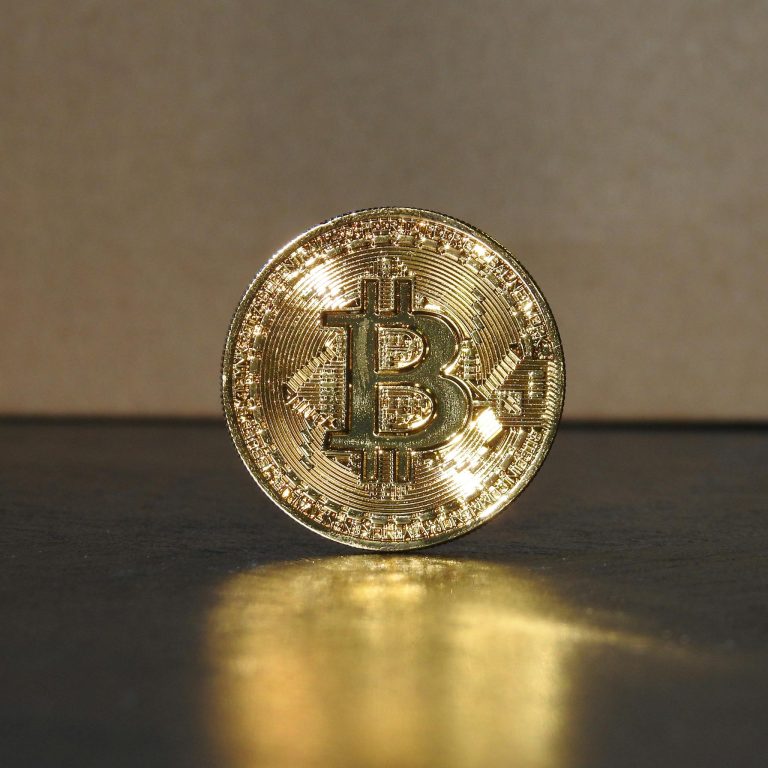 A shimmering golden Bitcoin coin standing upright on a reflective surface, illuminated by warm light.